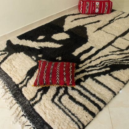 Beni Ourain abstract rug with black, white, and beige geometric pattern, fringed edges, displayed with a hat and red pouf