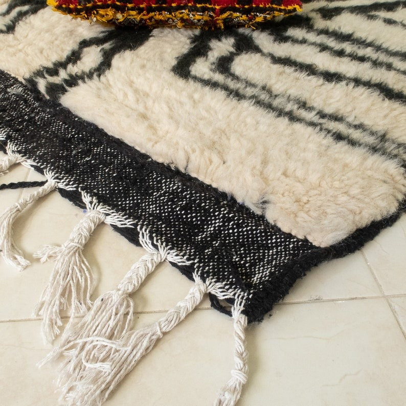 Beni Ourain abstract rug with black, white, and beige geometric pattern, fringed edges, displayed with a hat and red pouf