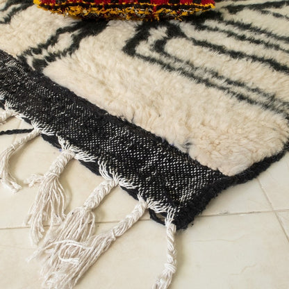 Beni Ourain abstract rug with black, white, and beige geometric pattern, fringed edges, displayed with a hat and red pouf