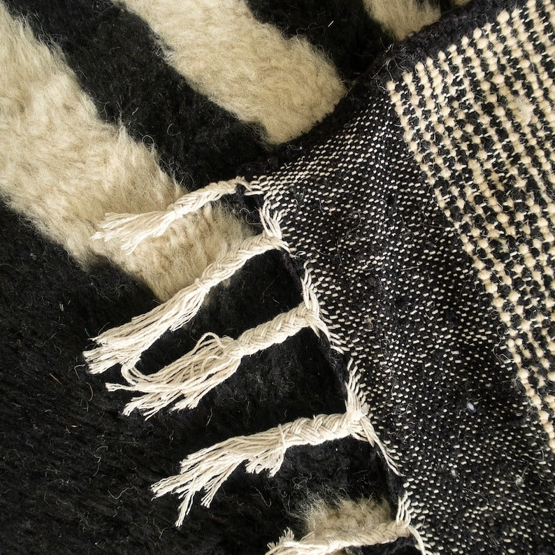 Beni Ourain abstract rug with black, white, and beige geometric pattern, fringed edges, displayed with a hat and red pouf.