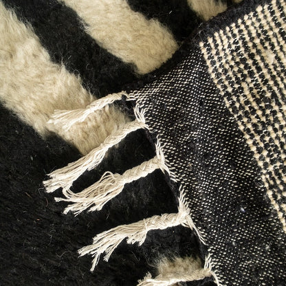 Beni Ourain abstract rug with black, white, and beige geometric pattern, fringed edges, displayed with a hat and red pouf.