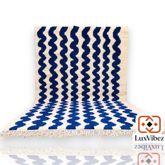 Blue Beni Ourain rug with white geometric patterns and fringed edges, displayed with a vase on a wooden floor.