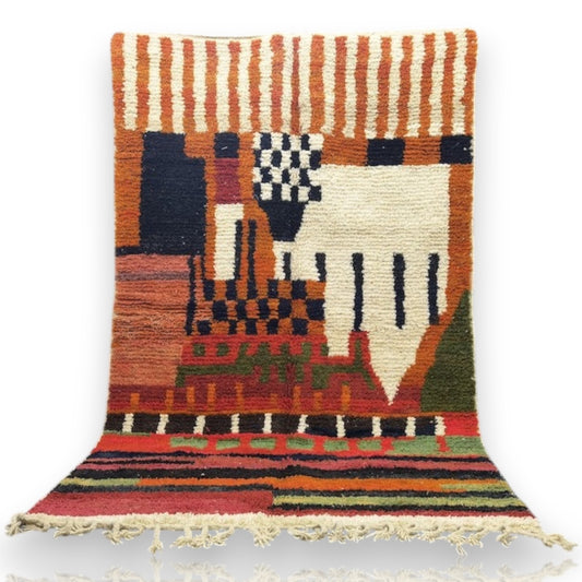 Orange Moroccan rug with multicolored geometric patterns and fringed edges, displayed against a curtained backdrop.