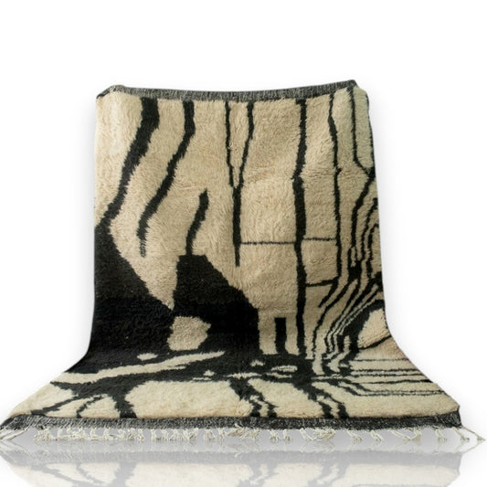 Beni Ourain abstract rug with black, white, and beige geometric pattern, fringed edges, displayed with a hat and red pouf